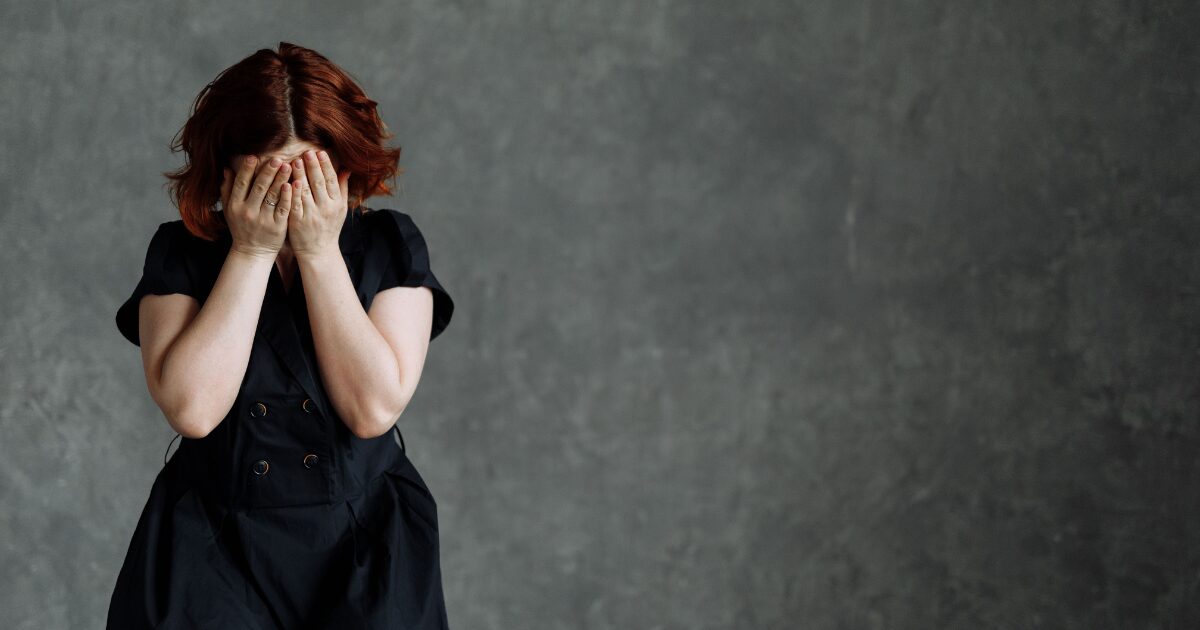 Woman with red hair in a black dress covers her face with both hands, standing against a gray textured wall, conveying distress or sadness.