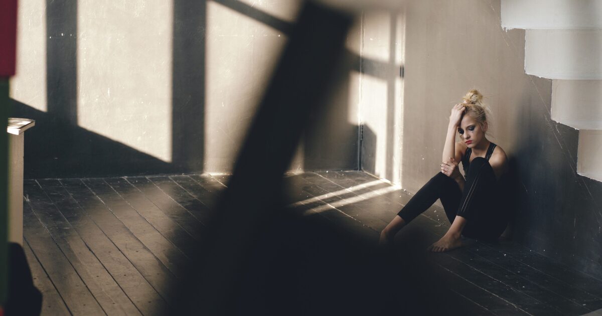 A woman sits on a dark wooden floor in a corner, leaning against the wall with her hand in her hair, looking contemplative in a dimly lit room.